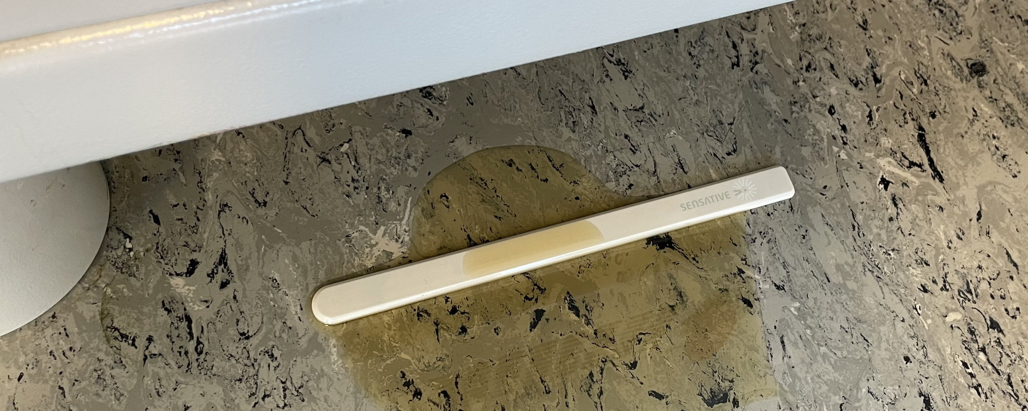 A Strips MS +Oil sensor lying on a workshop floor in a spreading pool of hydraulic oil, the oil drawn visibly into the absorber pad along part of the strip